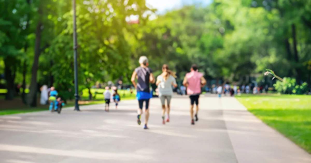 People jogging and cycling on a wide park path surrounded by trees.